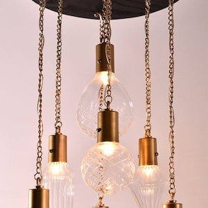 Puede incluir: Una lámpara de araña moderna con seis lámparas de vidrio transparente de varias formas, suspendidas de una base circular de madera oscura mediante cadenas doradas. Cada lámpara tiene una tapa de metal dorado.