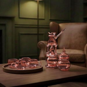 May include: A pink glass tea set on a wooden table. The set includes a teapot, a covered jar, and several teacups on a tray. The background features a leather armchair and a dark green wall.