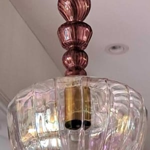 Könnte beinhalten: Eine Hängeleuchte mit einem transparenten, irisierenden Glasschirm und einem dekorativen, gestapelten, malvenfarbenen Glasstiel. Der Schirm hat ein geripptes, geschliffenes Glasdesign. Die Leuchte ist an einem weißen Kabel aufgehängt.