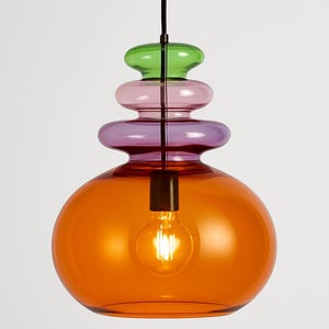 Könnte beinhalten: Moderne Pendelleuchte mit geschichtetem Design. Die Leuchte hat unten eine große orangefarbene Glaskugel, die von gestapelten Abschnitten aus lila, rosa und grünem Glas gekrönt wird. Im Inneren der orangefarbenen Kugel ist eine einzelne Glühbirne sichtbar.