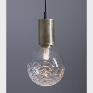 Puede incluir: Una lámpara colgante con un globo de vidrio transparente con un patrón de diamantes. La bombilla iluminada está conectada a un accesorio cilíndrico de color latón y un cable negro. La lámpara cuelga sobre un fondo gris.