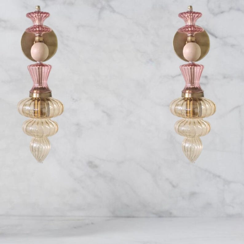 May include: Two decorative wall sconces with a vintage design. Each sconce features a pink, gold, and clear glass construction. The sconces have a layered, bulbous shape with a round gold backing and a pink top. The sconces are mounted on a white marble wall.