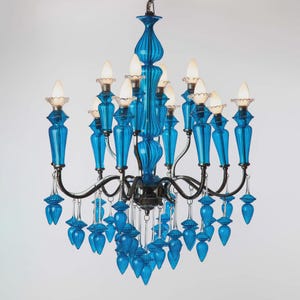 Puede incluir: Una lámpara de araña azul vibrante con múltiples accesorios de iluminación. La lámpara de araña presenta elementos azules ornamentados, similares al vidrio, y una estructura de metal oscuro. El diseño incluye adornos colgantes azules en forma de lágrima, creando una lámpara lujosa y decorativa.