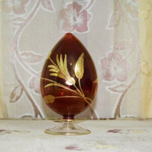 May include: A decorative, egg-shaped glass ornament in a rich, reddish-brown hue. The egg is adorned with gold-colored floral designs and rests on a clear, stemmed base with a gold rim. The background features a floral patterned curtain.