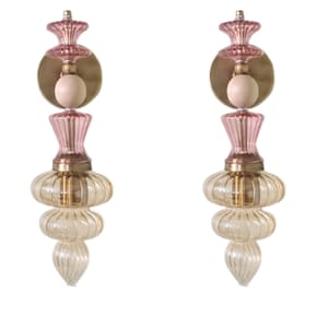 May include: Two decorative glass sconces with a layered design. Each sconce features a pink top, a brass-colored circular plate, and a light bulb. The lower section has a clear, ribbed glass design with a bulbous shape. The sconces are designed to be mounted on a wall.