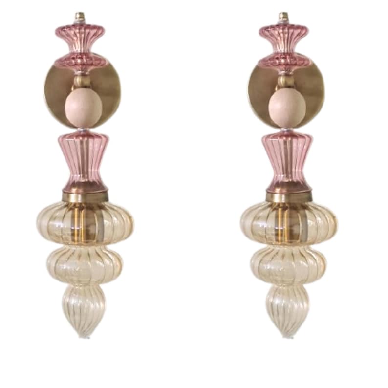May include: Two decorative glass sconces with a layered design. Each sconce features a pink top, a brass-colored circular plate, and a light bulb. The lower section has a clear, ribbed glass design with a bulbous shape. The sconces are designed to be mounted on a wall.