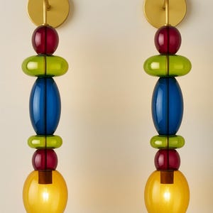 Può includere: Due applique da parete con un design colorato di perline di vetro impilate. Ogni applique presenta un supporto a parete circolare color oro, una sfera rossa, un ovale verde, un ovale blu e un apparecchio di illuminazione sferico giallo.