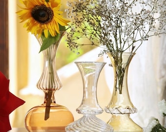 Op de afbeelding: Drie glazen vazen van verschillende vormen en maten. Eén vaas bevat een zonnebloem, een andere witte bloemen en de derde is leeg. De vazen zijn helder en amberkleurig, met decoratieve bodems en randen. De achtergrond is wazig.
