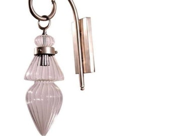 Può includere: Applique da parete con paralume in vetro trasparente a coste e finitura in metallo spazzolato. L'apparecchio ha un braccio curvo e un anello decorativo nella parte superiore. Il paralume in vetro ha la forma di una goccia.