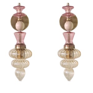 May include: Two decorative glass sconces with a layered design. Each sconce features a pink top, a brass-colored circular plate, and a light-colored bulb. The lower portion of the sconce has a clear, ribbed glass design.