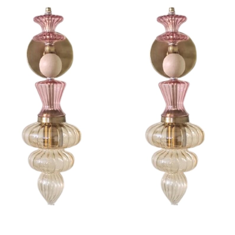May include: Two decorative glass sconces with a layered design. Each sconce features a pink top, a brass-colored circular plate, and a light-colored bulb. The lower portion of the sconce has a clear, ribbed glass design.