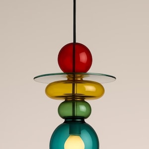 Puede incluir: Una moderna lámpara colgante con un diseño en capas. Cuenta con una esfera roja, un disco transparente, un óvalo amarillo, una esfera verde y una base turquesa. La bombilla es visible dentro de la base turquesa. La lámpara está suspendida de un soporte de techo dorado.