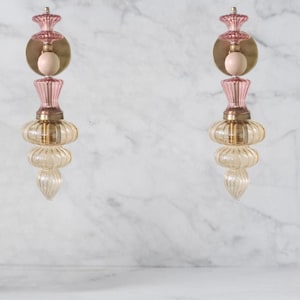 May include: Two decorative wall sconces. Each sconce features a pink, gold, and clear glass design. The sconces have a round gold base, a pink top, and a clear, ribbed glass bottom. The sconces are mounted on a white marble wall.
