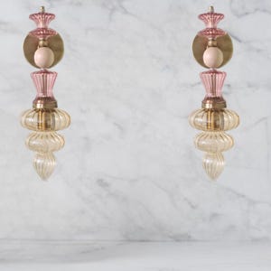 May include: Two decorative wall sconces. Each sconce features a pink, gold, and clear glass design. The sconces have a round gold base, a pink top, and a clear, ribbed glass bottom. The sconces are mounted on a white marble wall.