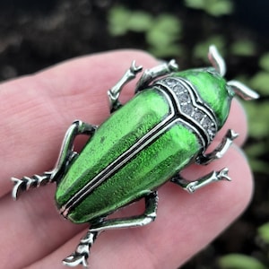 May include: A green beetle-shaped brooch with silver-colored legs and accents. The beetle's body is a vibrant green with a metallic sheen. The head features a decorative silver band with small, clear stones. The brooch is a unique accessory.