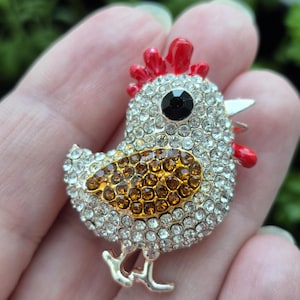 May include: A rhinestone chicken brooch with a red comb and wattle, a black eye, and a gold and brown belly. The brooch is made of silver-colored metal and is a decorative accessory.