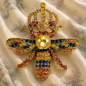 May include: A gold bee brooch with a crown, embellished with yellow, orange, blue, and red crystals. The brooch has a large yellow square crystal at its centre, set against a patterned fabric background.