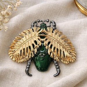 May include: A gold-toned metal insect brooch with green and black accents. The brooch features detailed gold leaf-shaped wings, a green gemstone body, and black legs and antennae. A unique and stylish accessory.