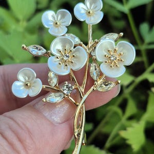 May include: A gold-tone metal brooch shaped like a bouquet of flowers. The piece features five white, petal-shaped components with gold centers, accented by clear, marquise-cut crystals arranged as leaves and small round stones on the stems.