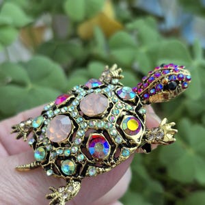 May include: A gold-coloured turtle brooch with an intricate design. The brooch has a shell decorated with iridescent and pink gemstones, and the head and feet are also embellished with smaller crystals. The brooch is held against a blurred green background.