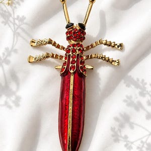 May include: A gold-toned insect brooch featuring a long, smooth red enamel body. The head and thorax are encrusted with small, sparkling red rhinestones, complemented by two black stone eyes and two slender antennae. It has four textured gold legs.