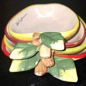 May include: A set of ceramic measuring cups in the shape of apples. The cups are stacked and feature a yellow, red, and green color scheme. The set includes a decorative leaf and stem design.