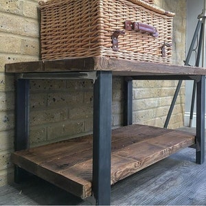 May include: Two woven wicker picnic baskets with leather straps and handles are stacked on a rustic wooden table with black metal legs. The baskets are light brown and have a rectangular shape. The table has a lower shelf.