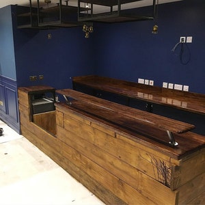 Puede incluir: Una barra de madera hecha a medida con una encimera oscura y una base de madera clara y tosca. La barra cuenta con una secci&oacute;n empotrada y est&aacute; colocada contra una pared azul oscuro. Encima, hay estanter&iacute;as met&aacute;licas y luces colgantes.