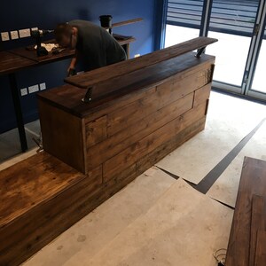 Puede incluir: Una barra de madera en construcci&oacute;n, con una mancha oscura. La barra tiene una larga encimera y un escal&oacute;n inferior. Las paredes est&aacute;n pintadas de azul oscuro y el suelo es de hormig&oacute;n. Una persona est&aacute; trabajando en la barra.