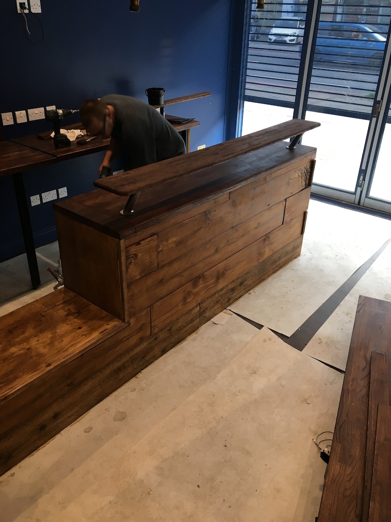 Puede incluir: Una barra de madera en construcci&oacute;n, con una mancha oscura. La barra tiene una larga encimera y un escal&oacute;n inferior. Las paredes est&aacute;n pintadas de azul oscuro y el suelo es de hormig&oacute;n. Una persona est&aacute; trabajando en la barra.
