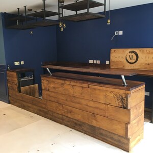 Puede incluir: Un bar de madera hecho a medida con un tinte oscuro, con una encimera elevada y un &aacute;rea de servicio inferior. El bar tiene un dise&ntilde;o r&uacute;stico con vetas de madera visibles y un elemento decorativo. Arriba, hay estantes con marco de metal y luces colgantes.
