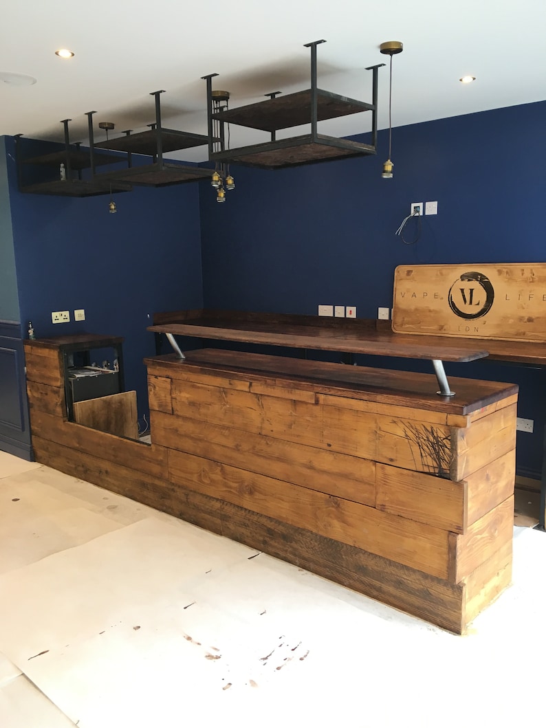 Puede incluir: Un bar de madera hecho a medida con un tinte oscuro, con una encimera elevada y un &aacute;rea de servicio inferior. El bar tiene un dise&ntilde;o r&uacute;stico con vetas de madera visibles y un elemento decorativo. Arriba, hay estantes con marco de metal y luces colgantes.
