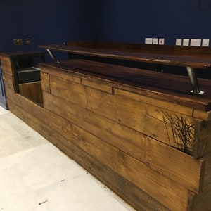 Puede incluir: Una barra de madera hecha a medida con un acabado te&ntilde;ido oscuro. La barra presenta un dise&ntilde;o de varios niveles con un &aacute;rea de servicio y una barra elevada. La madera tiene una apariencia r&uacute;stica y desgastada. La barra est&aacute; contra una pared azul oscuro.