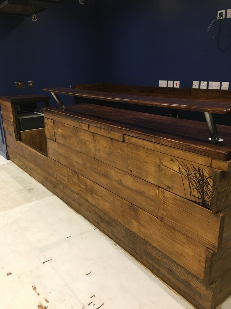 Puede incluir: Una barra de madera hecha a medida con un acabado te&ntilde;ido oscuro. La barra presenta un dise&ntilde;o de varios niveles con un &aacute;rea de servicio y una barra elevada. La madera tiene una apariencia r&uacute;stica y desgastada. La barra est&aacute; contra una pared azul oscuro.