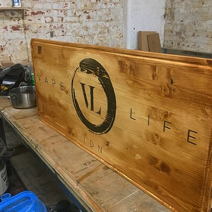 Puede incluir: Un letrero de madera rectangular grande con las palabras "VAPE LIFE LDN" en letras negras. Las letras "VL" est&aacute;n dentro de un c&iacute;rculo negro. La madera tiene una mancha marr&oacute;n c&aacute;lida.