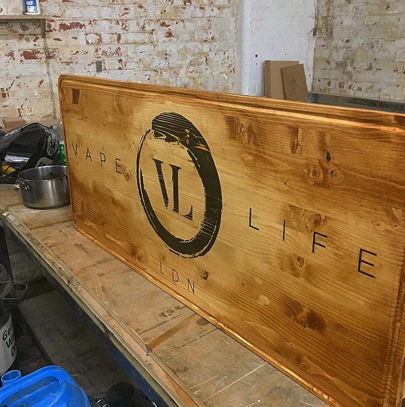 Puede incluir: Un letrero de madera rectangular grande con las palabras "VAPE LIFE LDN" en letras negras. Las letras "VL" est&aacute;n dentro de un c&iacute;rculo negro. La madera tiene una mancha marr&oacute;n c&aacute;lida.