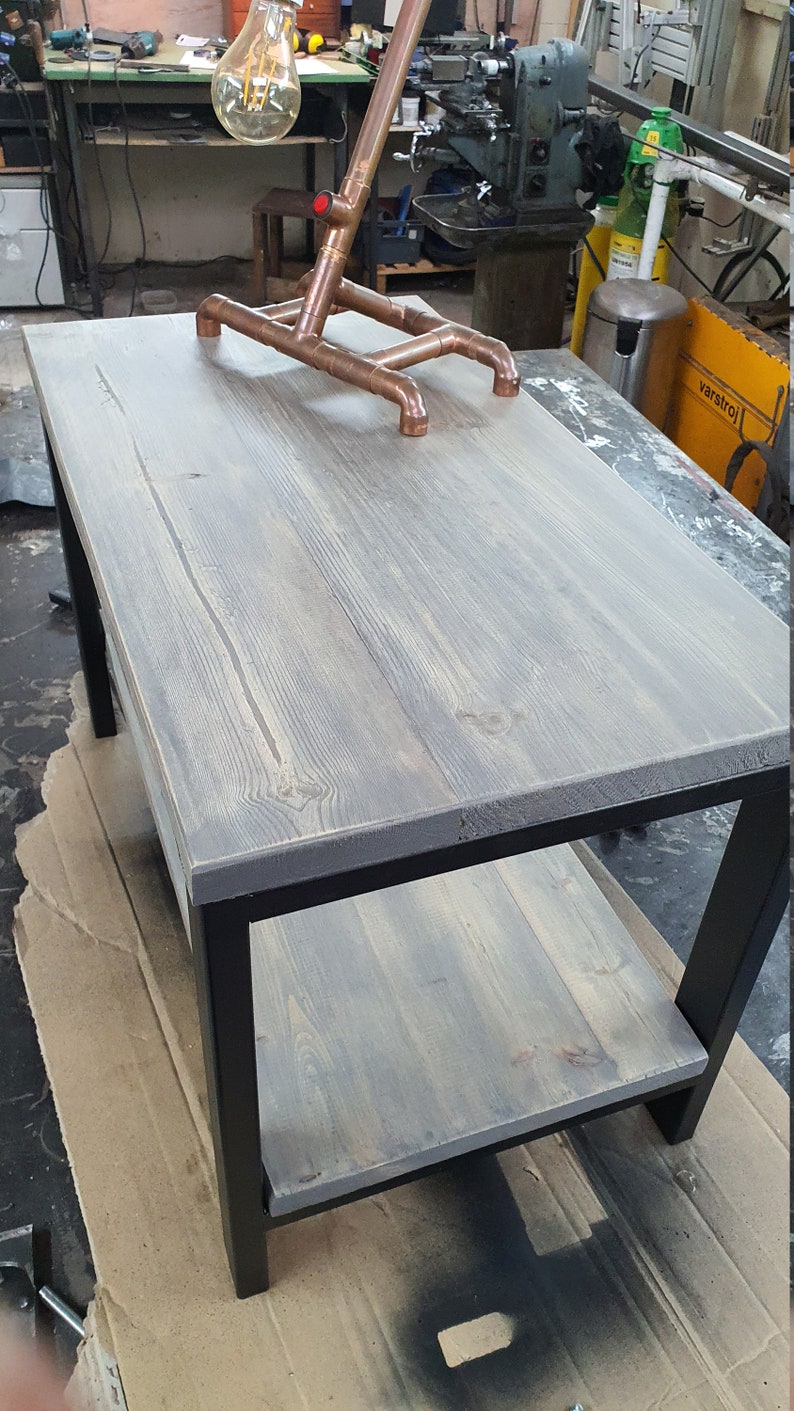 May include: A gray wooden coffee table with a black metal frame and a lower shelf. A copper pipe lamp with a large bulb sits on the table. The table is approximately 48 inches long and 24 inches wide.