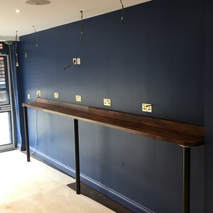 Puede incluir: Vista interior de una habitaci&oacute;n en construcci&oacute;n. Una larga encimera de madera oscura est&aacute; sostenida por patas de metal negro contra una pared azul oscuro. Se han instalado varios enchufes el&eacute;ctricos. Los cables expuestos cuelgan del techo y el suelo est&aacute; cubierto con material protector.