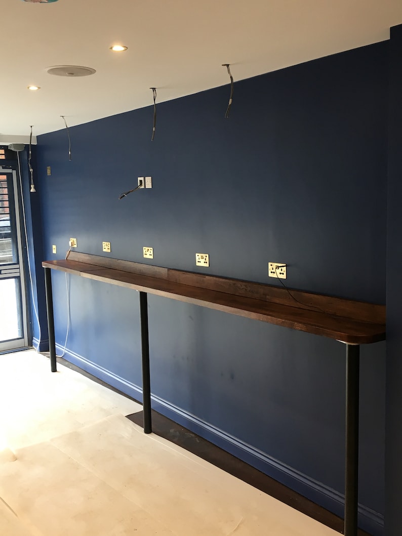 Puede incluir: Vista interior de una habitaci&oacute;n en construcci&oacute;n. Una larga encimera de madera oscura est&aacute; sostenida por patas de metal negro contra una pared azul oscuro. Se han instalado varios enchufes el&eacute;ctricos. Los cables expuestos cuelgan del techo y el suelo est&aacute; cubierto con material protector.