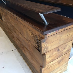 Puede incluir: Una barra de madera hecha a medida con una encimera te&ntilde;ida oscura y una base de madera m&aacute;s clara. La barra cuenta con una encimera elevada sostenida por patas de metal pulido. La veta de la madera es visible y la barra parece de nueva construcci&oacute;n.
