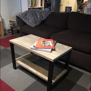 May include: A two-tiered coffee table with a light wood top and shelf, supported by black metal legs. Books are stacked on the top surface. The table is approximately 36 inches long, 18 inches wide, and 18 inches high.