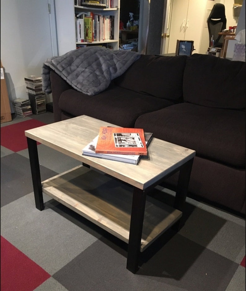 May include: A two-tiered coffee table with a light wood top and shelf, supported by black metal legs. Books are stacked on the top surface. The table is approximately 36 inches long, 18 inches wide, and 18 inches high.