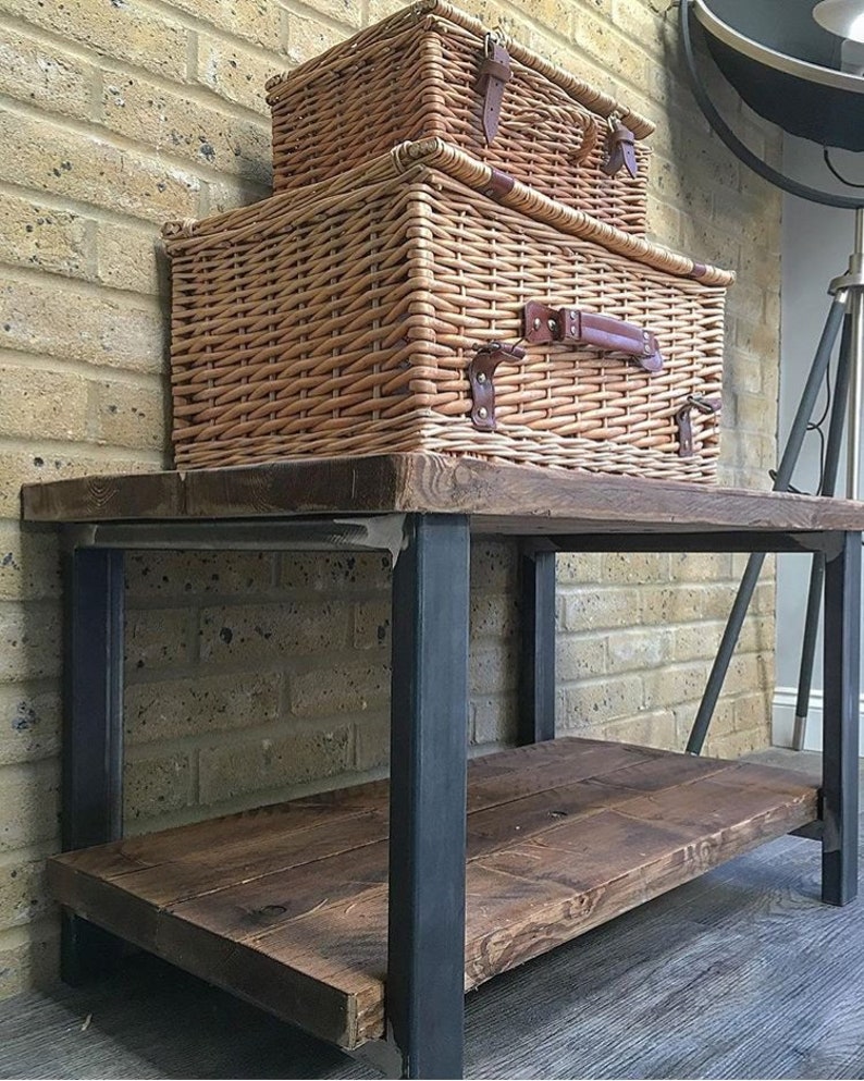 May include: Two woven wicker picnic baskets with leather straps and handles are stacked on a rustic wooden table with black metal legs. The baskets are light brown and have a rectangular shape. The table has a lower shelf.