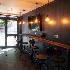 Puede incluir: Vista interior de una zona de bar con una pared azul oscuro, una encimera de madera y varias banquetas de bar de metal y madera. Las bombillas de estilo Edison cuelgan del techo. Un televisor grande est&aacute; montado en la pared. Se ve una puerta con persianas.