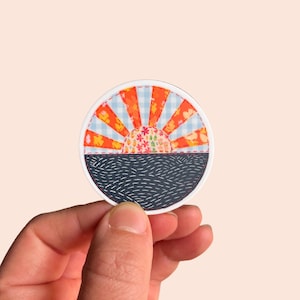 May include: A round sticker with a colorful patchwork design of a sunset over a dark blue sea. The sticker is white with a red, orange, yellow, and blue design.