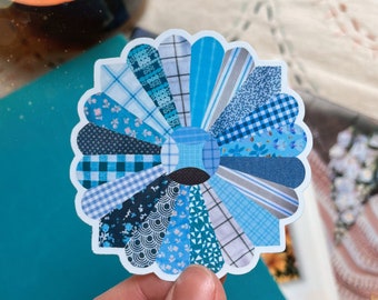 May include: A blue and white patchwork flower sticker with a circular design. The sticker is made up of different blue and white patterned fabrics.