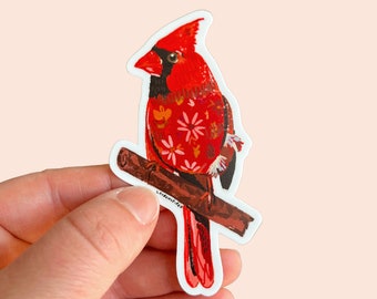 Può includere: Un adesivo con un cardinale rosso con un motivo floreale sul petto. L'uccello è appollaiato su un ramo con una texture marrone e bianca. L'adesivo è su uno sfondo bianco.