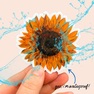 Peut inclure: Un autocollant imperm&eacute;able repr&eacute;sentant un tournesol &agrave; l'aquarelle avec des p&eacute;tales jaunes et un centre brun fonc&eacute;. L'autocollant a une bordure blanche et est &eacute;clabouss&eacute; de gouttelettes d'eau bleues. Le texte sur l'image indique : "yes, I'm waterproof!"