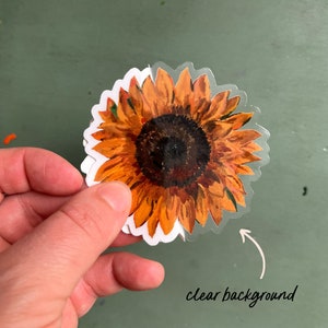 Peut inclure: Un autocollant transparent repr&eacute;sentant un tournesol &agrave; l'aquarelle avec des p&eacute;tales orange et jaunes et un centre brun fonc&eacute;. L'autocollant a une bordure blanche et un fond transparent. Le texte "clear background" est visible.