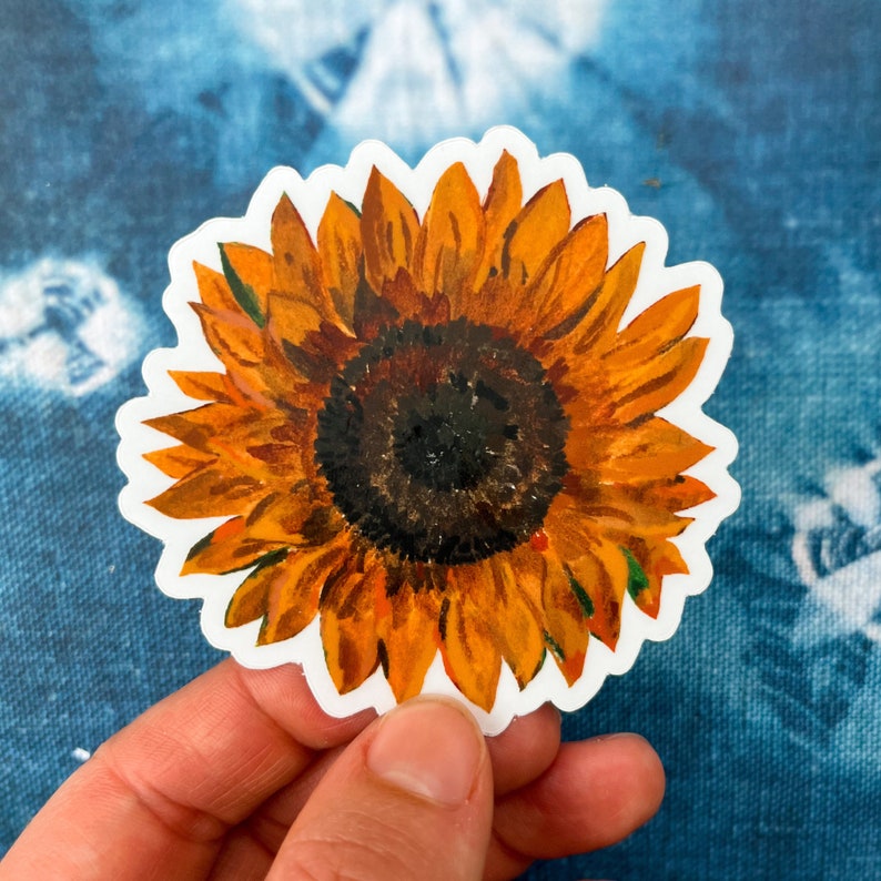 Peut inclure: Un autocollant d&eacute;coup&eacute; repr&eacute;sentant une peinture &agrave; l'aquarelle d'un tournesol. Le tournesol a des p&eacute;tales jaune-orang&eacute; vif et un centre brun fonc&eacute;. L'autocollant est tenu dans une main sur un fond bleu.